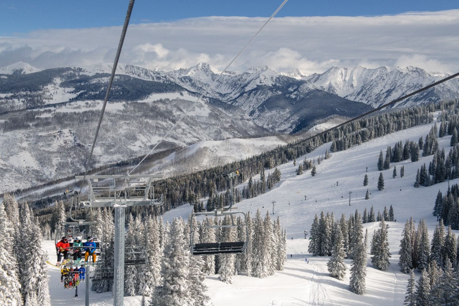 Skiers ride a chairlift above snow-covered runs and pine trees, with snowcapped Colorado peaks beyond