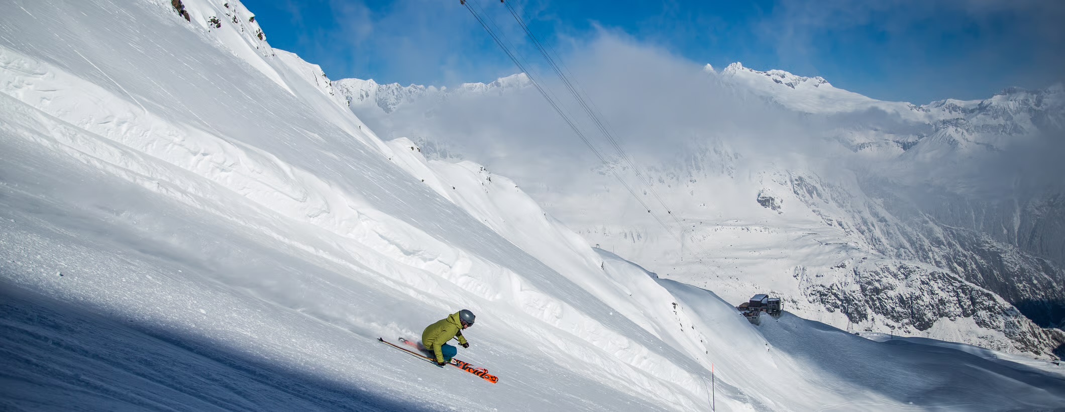 A skier carving down the steep, sunlit Russi Run on the Gemsstock, with cloud-wrapped peaks beyond
