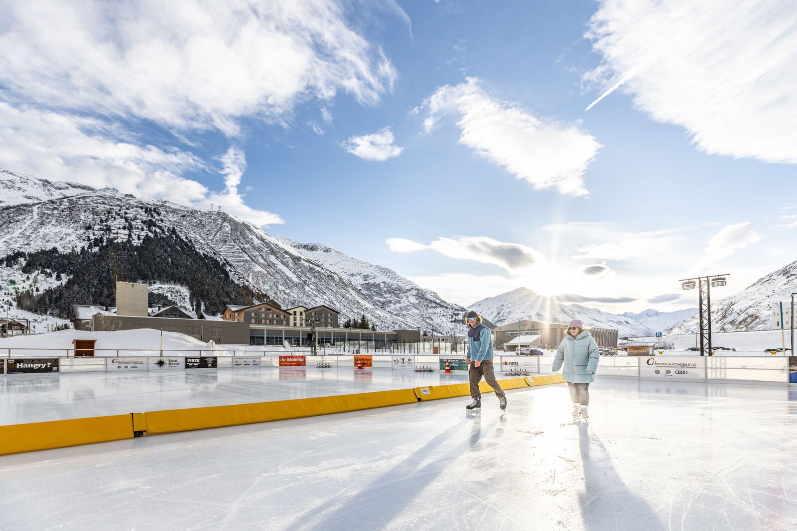 Two skaters gliding across the outdoor ice rink in Andermatt on a sunny winter day