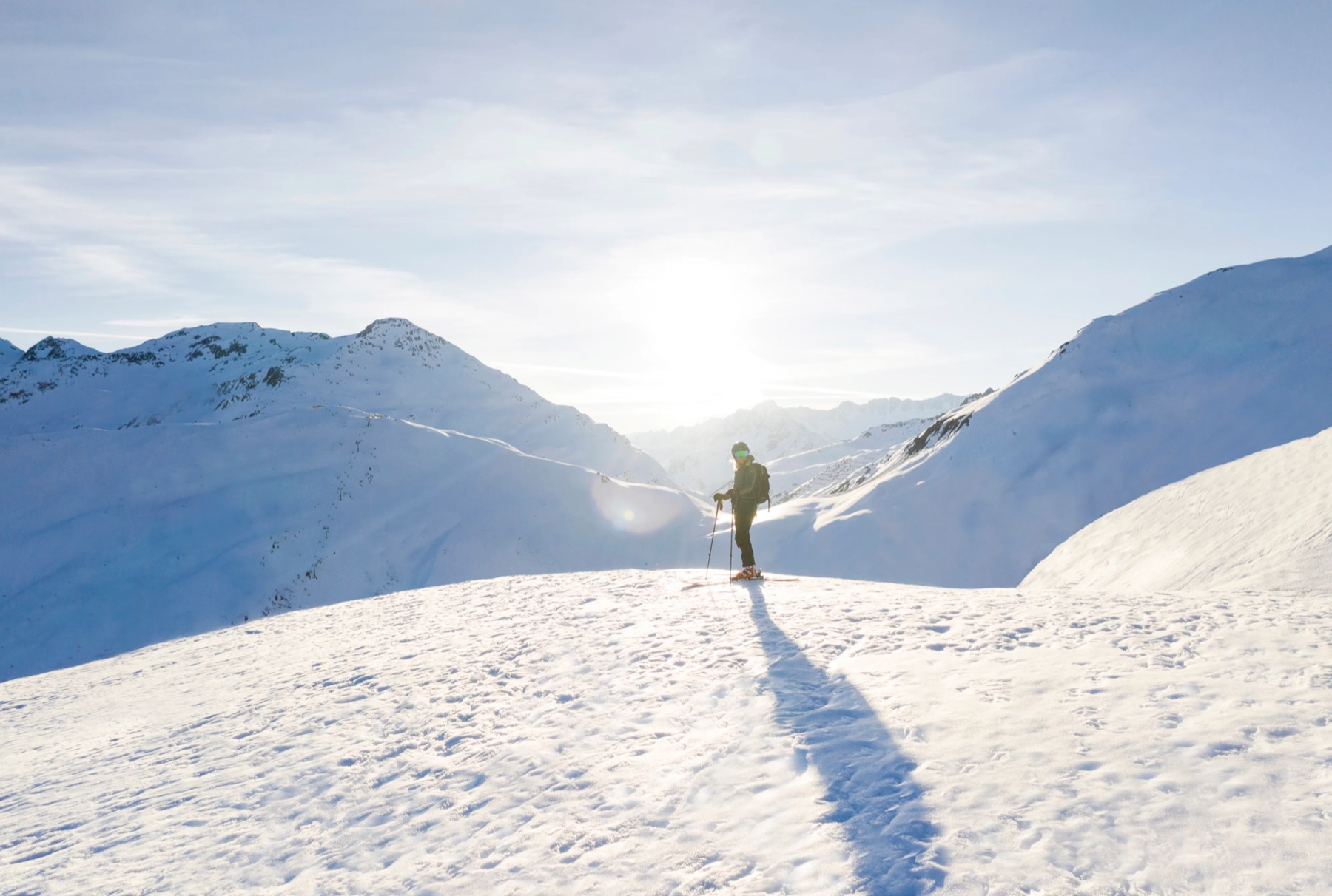 A skier pausing on a sunny snowfield above a high Alpine valley, with snowy peaks all around