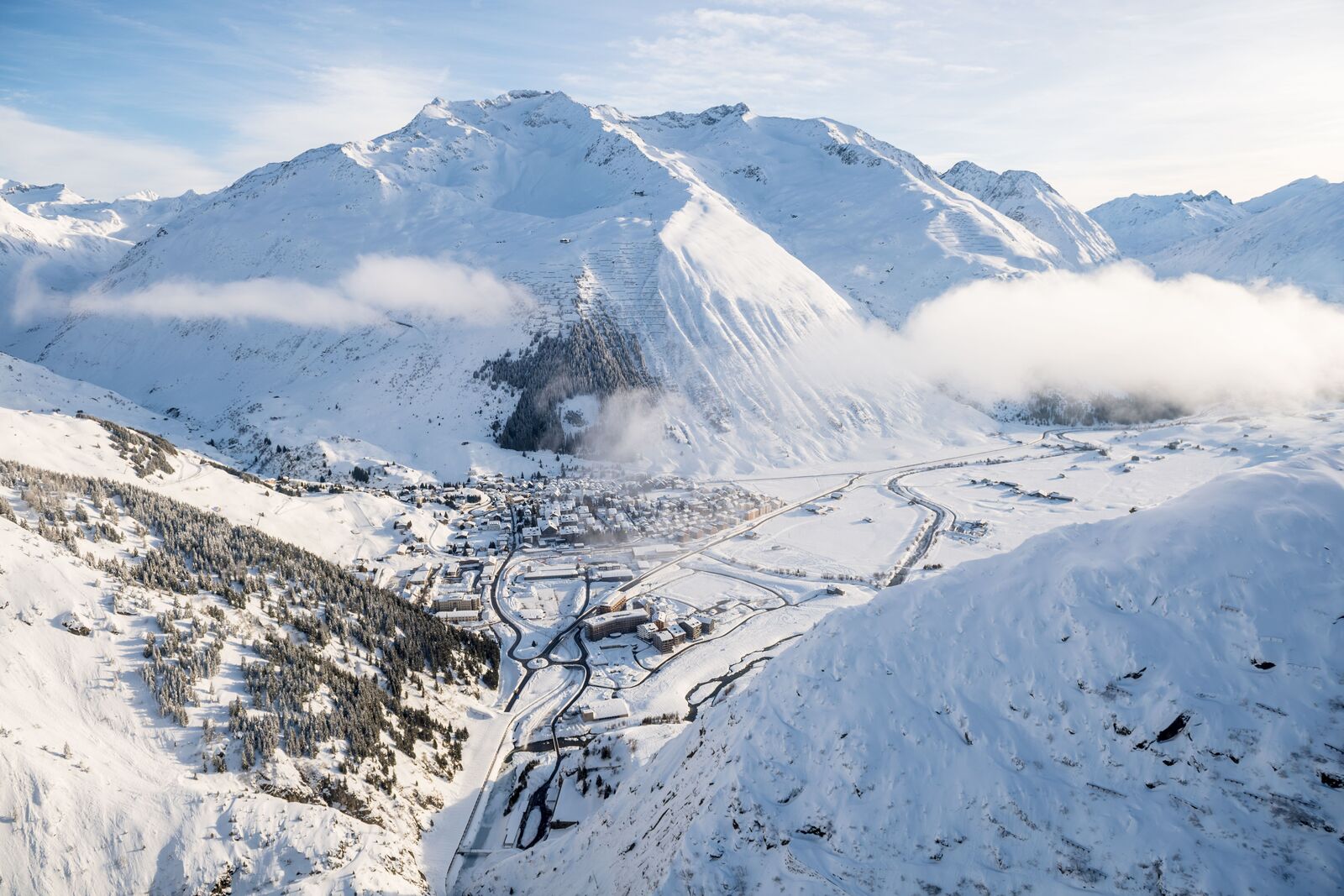 Andermatt seen from above, its snowy valley floor ringed by high white peaks and drifting cloud