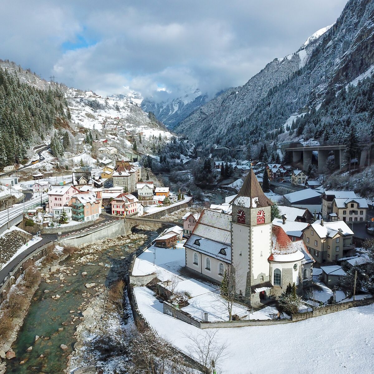The village of Gurtnellen and its church beside a mountain river, with a railway viaduct and snowy peaks behind