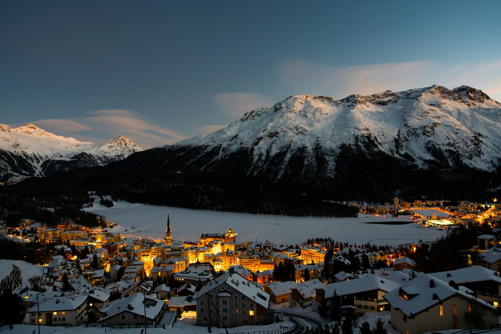 St. Moritz glowing at dusk beside its frozen lake, beneath snow-covered peaks