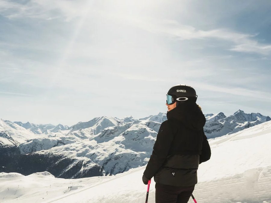 A skier pausing on a sunny ridge above a sweep of snowy Engadin peaks
