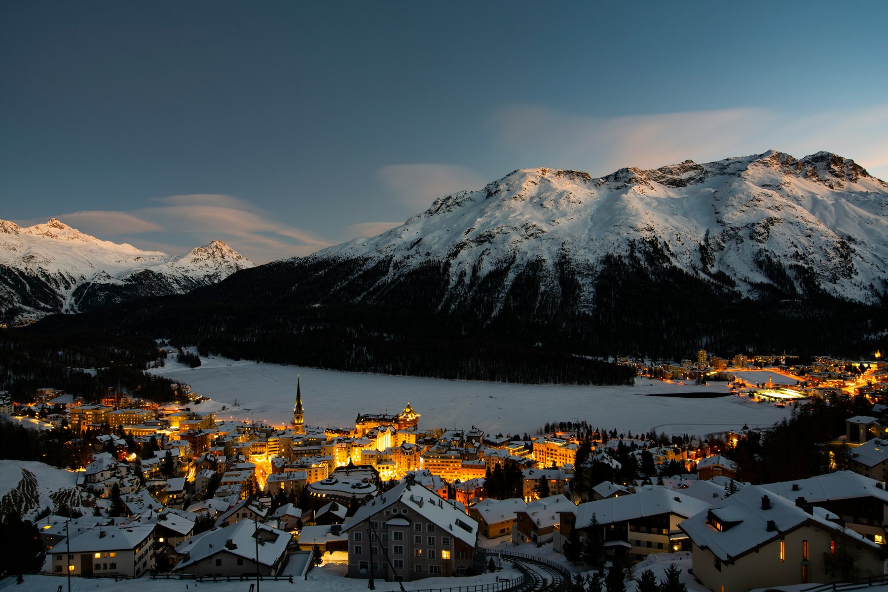 St. Moritz glowing at twilight above the frozen lake, with snowy peaks behind
