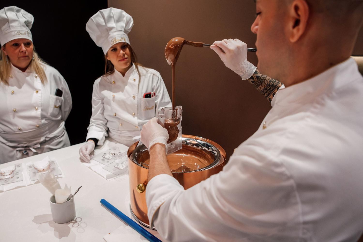 A Lindt master chocolatier ladling melted chocolate into a mold as guests in chef's whites look on