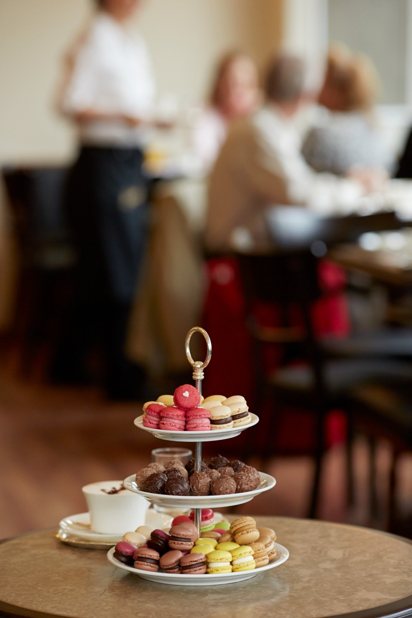 A tiered stand of Luxemburgerli and chocolate truffles beside a cappuccino at Confiserie Sprüngli