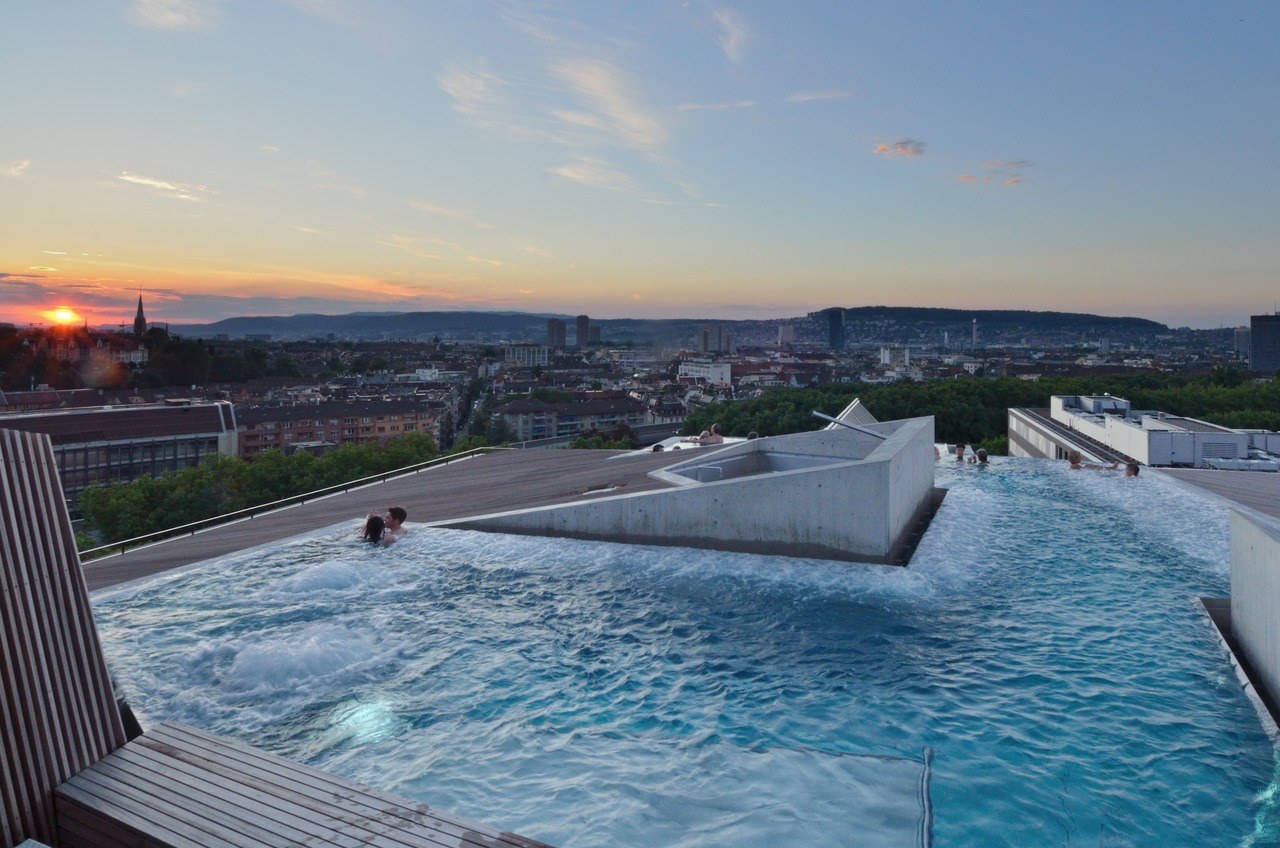 Bathers in the rooftop thermal pool at Thermalbad & Spa Zurich, with the city below at sunset